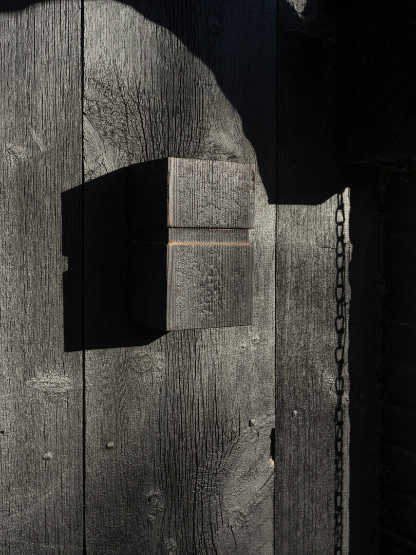 A rectangular wooden box mounted on a dark, weathered wooden wall with a chain hanging to the right, partially illuminated by sunlight.